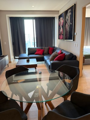 Een moderne woonkamer met een salontafel met glazen blad, zwarte banken met rode kussens en een groot raam met gordijnen.