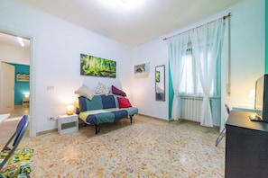 Un dormitorio con cama, mesita de noche, escritorio, silla, ventana con cortinas y un cuadro en la pared.