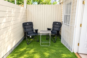 Eine Terrassengruppe im Freien mit zwei schwarzen Klappstühlen und einem kleinen Tisch, umgeben von einem weißen Zaun und einer weißen Tür mit Insektenschutzgitter.