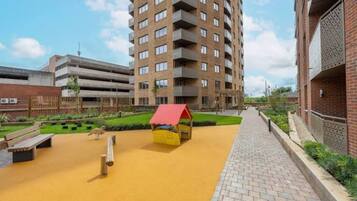 A multi-story residential building with balconies, a paved walkway, and a small play area with a red-roofed structure.