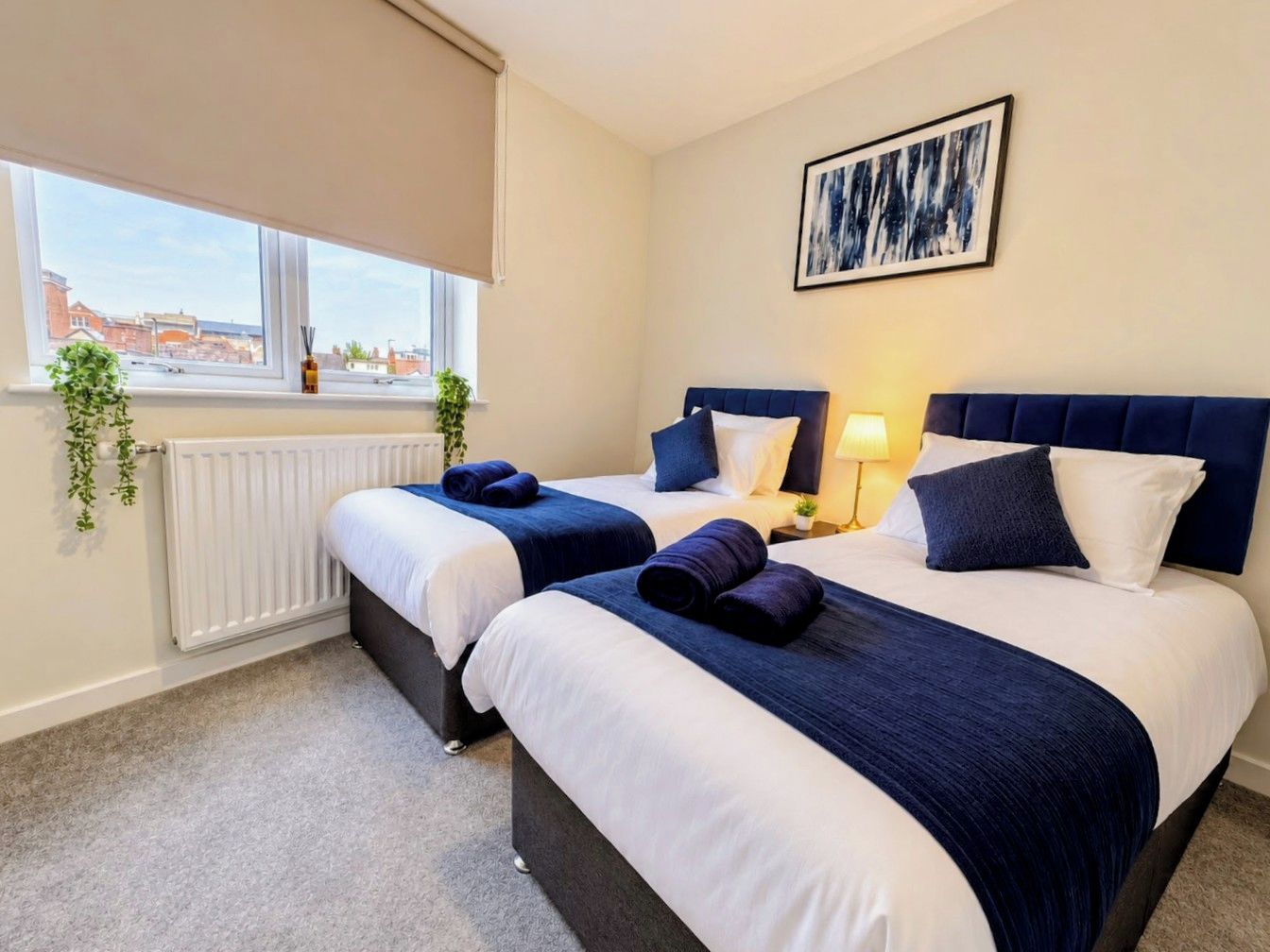 A hotel room with two beds, a radiator, a window with a view, and a framed picture on the wall.