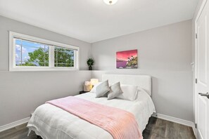 A bedroom with a bed, a window, a bedside table with a lamp, and a wall-mounted picture.
