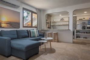 A modern living room with a blue sofa, a small round table, and a kitchen area with white cabinets and a water dispenser. - Casita Cuna (Puerto Morelos)
