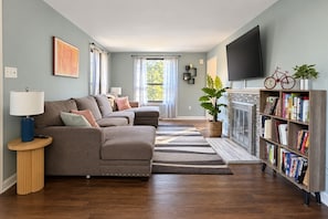 A living room with a sectional sofa, a coffee table, a bookshelf, a fireplace, and a television.