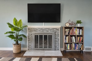 A living room with a fireplace, a flat-screen TV mounted above it, a bookshelf filled with books, a potted plant, and a bicycle.