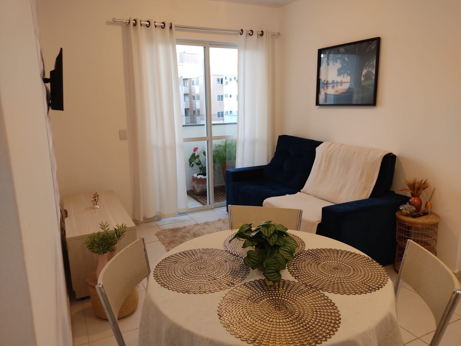 A living room with a sofa, a dining table with placemats, a small side table with a vase, and a sliding glass door leading to a balcony.