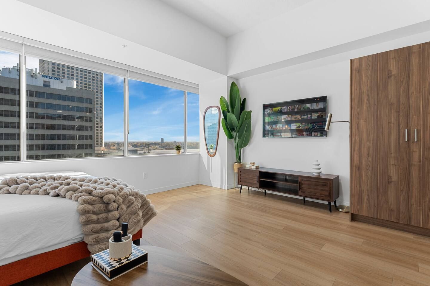 A modern bedroom with a large bed, a wooden dresser, a mirror, and a city view.