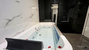 A modern bathroom with a marble wall, a bathtub with rose petals, and a glass shower enclosure. - Suisse Luxury Residence (Nallur)