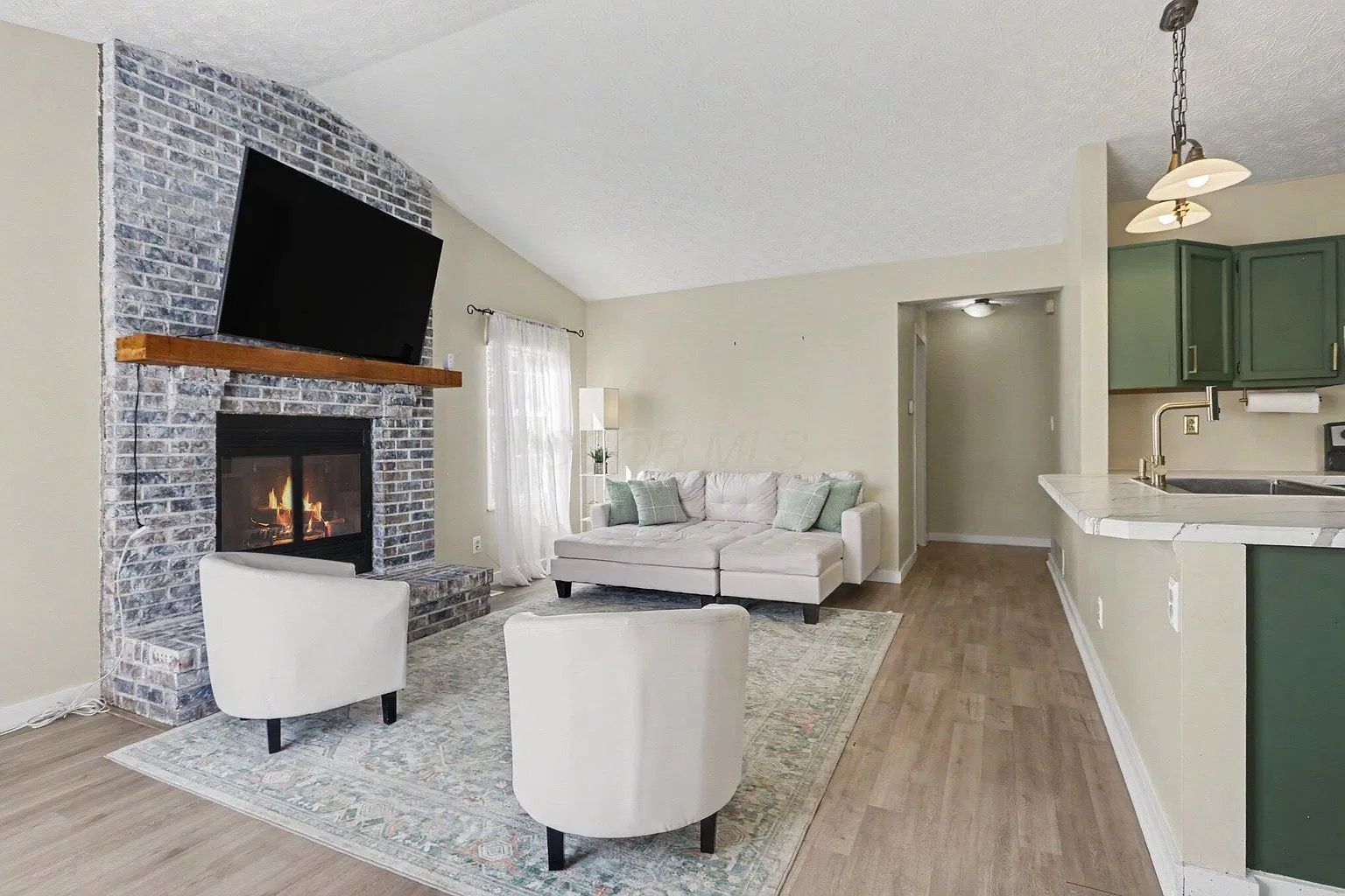 A living room with a fireplace, a sofa, two armchairs, and a kitchen area with green cabinets.