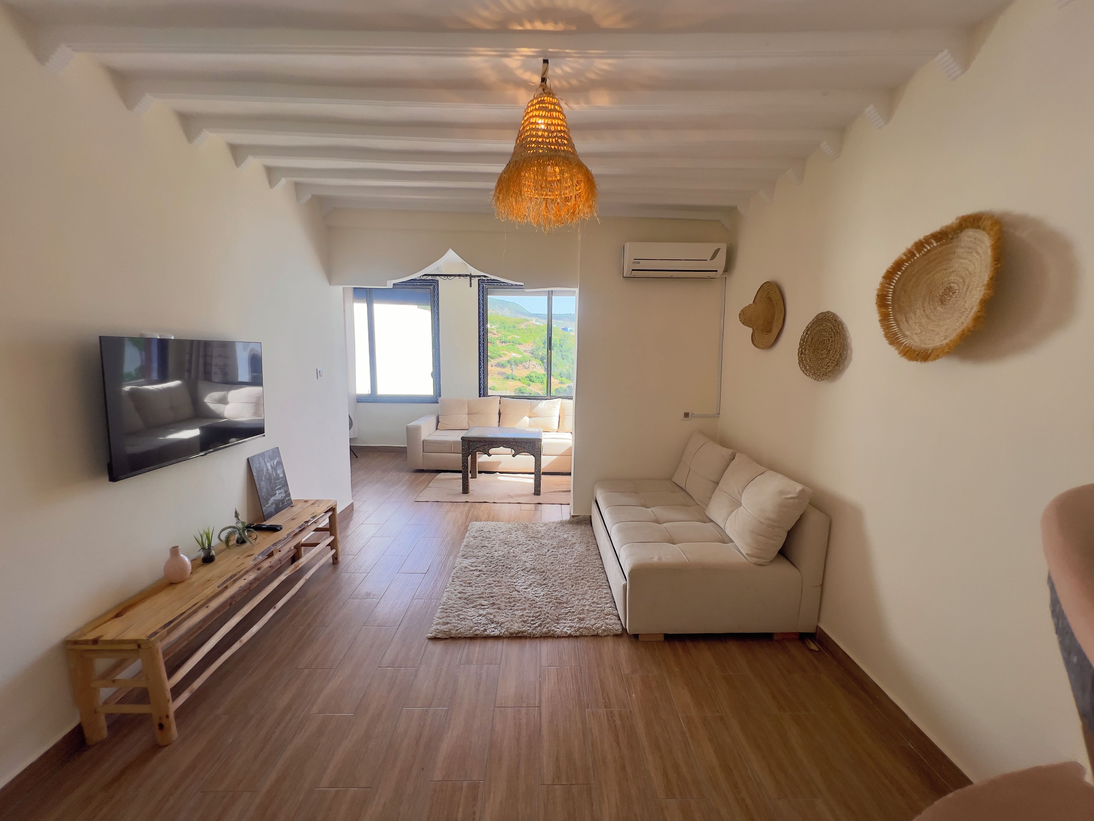 Un salón espacioso con una televisión de pantalla plana, un banco de madera, un sofá color beige y un ventanal con vistas al exterior.