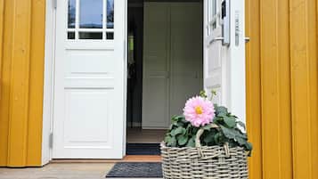 Una puerta de madera con marco blanco, una cesta con una flor rosa y una terraza de madera que conduce a la entrada.