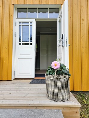 Una puerta de madera con marco blanco, una cesta con una flor rosa y una terraza de madera que conduce a la entrada.