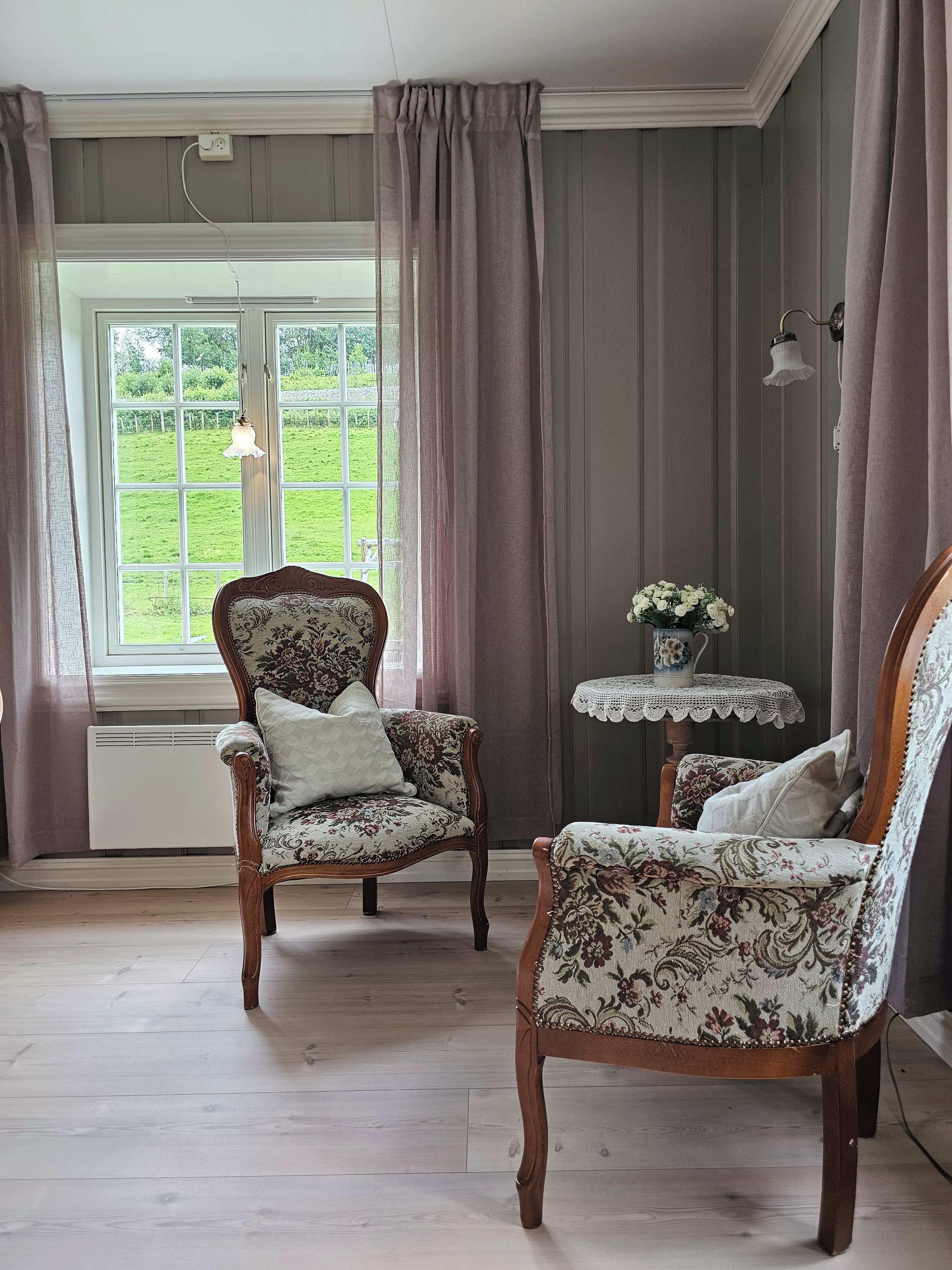 A room with two upholstered chairs, a small table with a vase of flowers, and a window with curtains.
