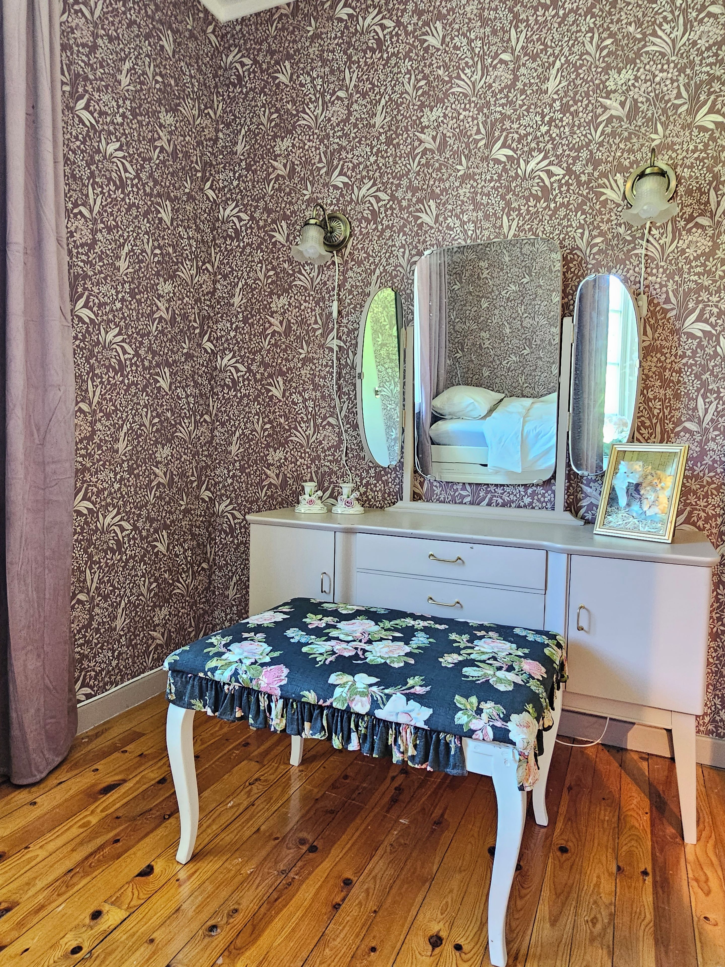 A bedroom with floral wallpaper, a bed with white bedding, a dresser with a mirror, and a bench with a floral cover.