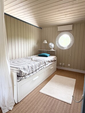 Ein Schlafzimmer mit einem Einzelbett, einem Nachttisch mit Lampe und Ventilator, einem Fenster und einem Teppichboden.