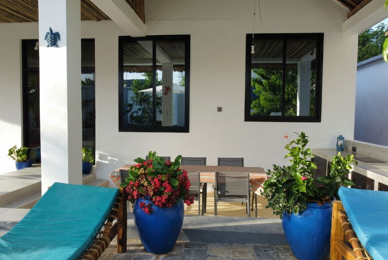 Un espacio de estar al aire libre techado con mesa, sillas y una silla de descanso, además de dos grandes macetas con plantas.