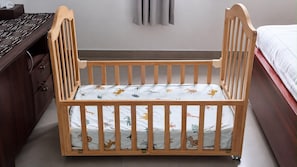 Una culla in legno con un materasso a motivi, situata in una camera da letto con una cassettiera in legno scuro e un letto con copripiumino bianco.