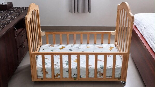 Un berceau en bois avec un matelas à motifs, placé dans une chambre où se trouvent une commode en bois foncé et un lit recouvert d’une couette blanche.