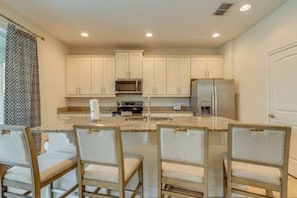 A kitchen with white cabinets, a stainless steel refrigerator, and a central island with bar stools. - ECR7391 - Family Luxury Retreat + Pool @Encore! (Kissimmee)