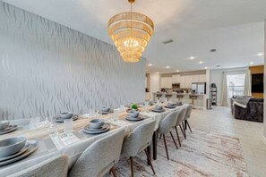 A dining area with a long table set for a meal, a chandelier, and a kitchen in the background. - ECR313 - Resort Style Mansion + Gyn/Spa/Theater! (Kissimmee)