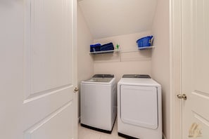 A laundry room with two front-loading washing machines and a shelf above holding folded towels and laundry baskets. - ECR456 - Luxury Resort Design Home @ Encore! (Kissimmee)