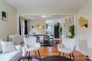 A modern living room with a sofa, two armchairs, a dining table with chairs, a radiator, and a kitchen in the background.