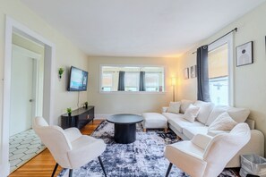 A modern living room with a sofa, chairs, a coffee table, a TV, and a window with blinds.