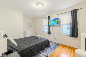A bedroom with a bed, a flat-screen TV, a bedside table, a window with curtains, and a radiator.