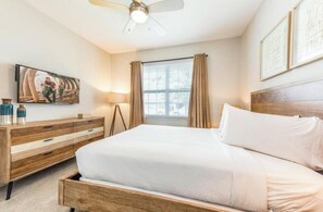 Une chambre d’hôtel avec un lit, une commode en bois, un ventilateur de plafond, une fenêtre avec des rideaux et des œuvres d’art accrochées au mur.
