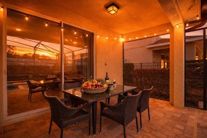 Eine Terrasse im Freien mit Glasveranda, Korbmöbeln, einem Tisch mit Früchten und einer Flasche sowie Lichterketten.