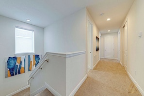 A hallway with a carpeted floor, white walls, a window with blinds, and a staircase with a handrail. - ECR7739 - Resort Style Home 10 min from Disney! (Kissimmee)