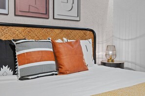 A neatly made bed with striped and solid pillows, a woven headboard, and a bedside lamp on a round table. - ECR3385 Only 13 Minutes from Disney | Pool & GYN (Kissimmee)