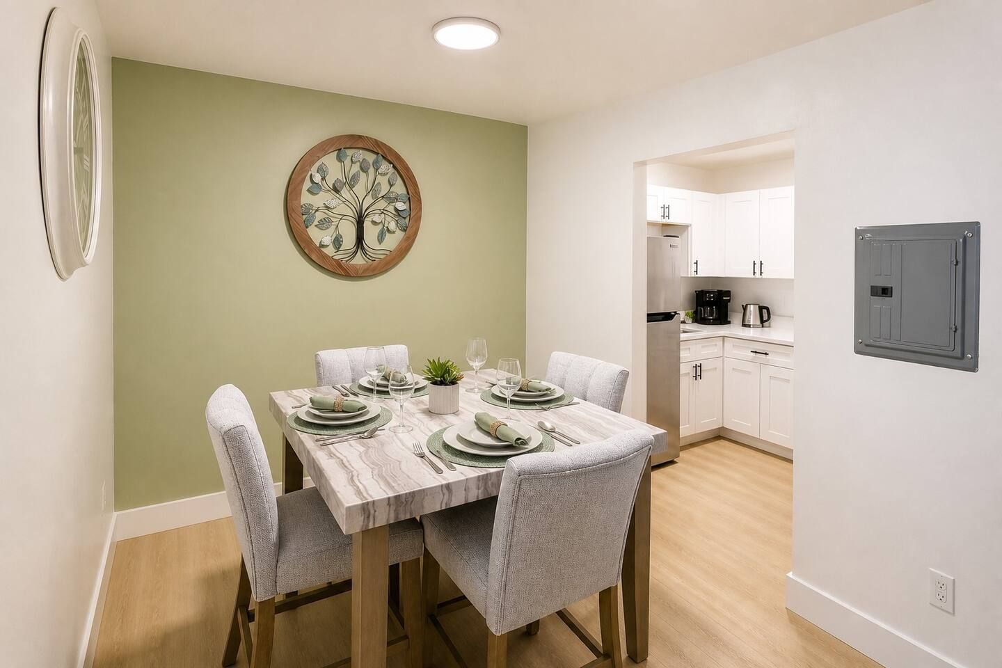 A dining area with a table set for four, chairs, a wall-mounted clock, and a kitchen visible in the background.