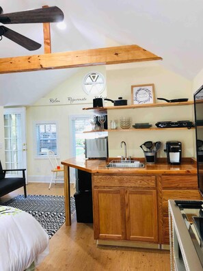 A compact kitchen with wooden cabinets, a built-in oven, a coffee maker, and a window with blinds. - Hollytree Cottage at Saluda River Farms (Greenville)