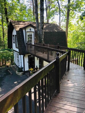A wooden deck with a railing, leading to a small white building with a dark roof, surrounded by trees. - Hollytree Cottage at Saluda River Farms (Greenville)