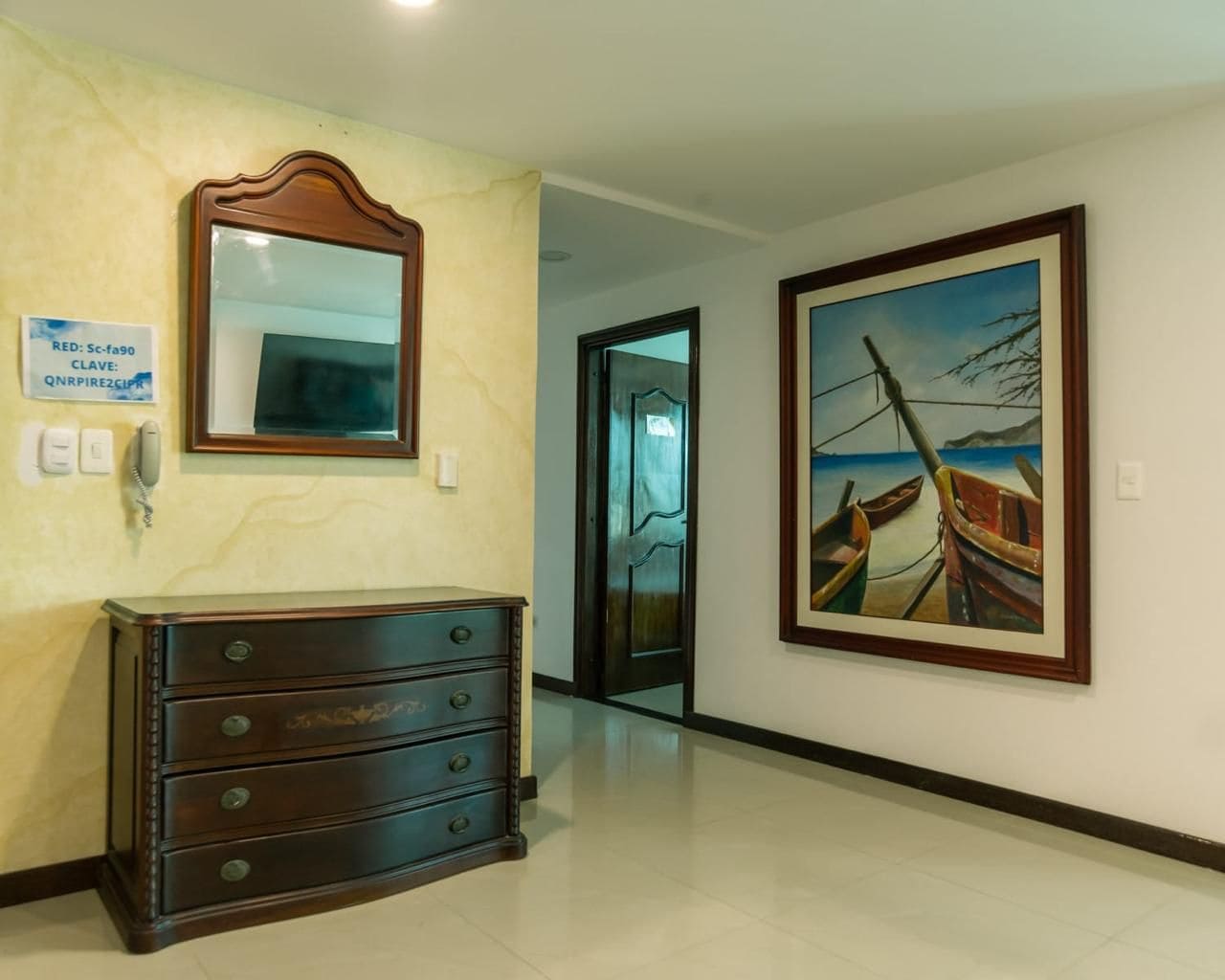 A hallway with a wooden dresser, a mirror, a painting of boats, and a door leading to another room.
