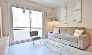 Ein modernes Wohnzimmer mit einem Glastisch, einem grauen Sessel, einem beigen Sofa mit Kissen und einer raumhohen Schiebetür zum Balkon.
