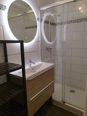 A modern bathroom with a glass-enclosed shower, a wooden vanity with a sink, and a round mirror above the vanity. - New Listing (Le Grau-du-Roi)