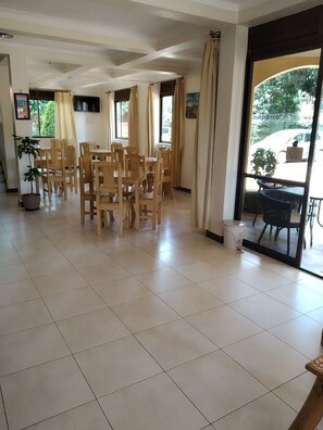 A spacious dining area with multiple wooden tables and chairs, a television mounted on the wall, and large windows with curtains. - The Dove's Nest Hotel Gulu (Gulu)