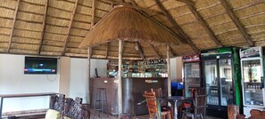 A bar area with a thatched roof, a TV mounted on the wall, and a refrigerator stocked with beverages. - The Dove's Nest Hotel Gulu (Gulu)