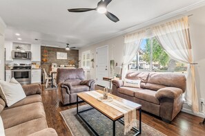 A modern living room with a sofa, armchair, and coffee table, a kitchen with appliances, and a dining area. - City of Trees House with Hot Tub & Game Room (West Sacramento)