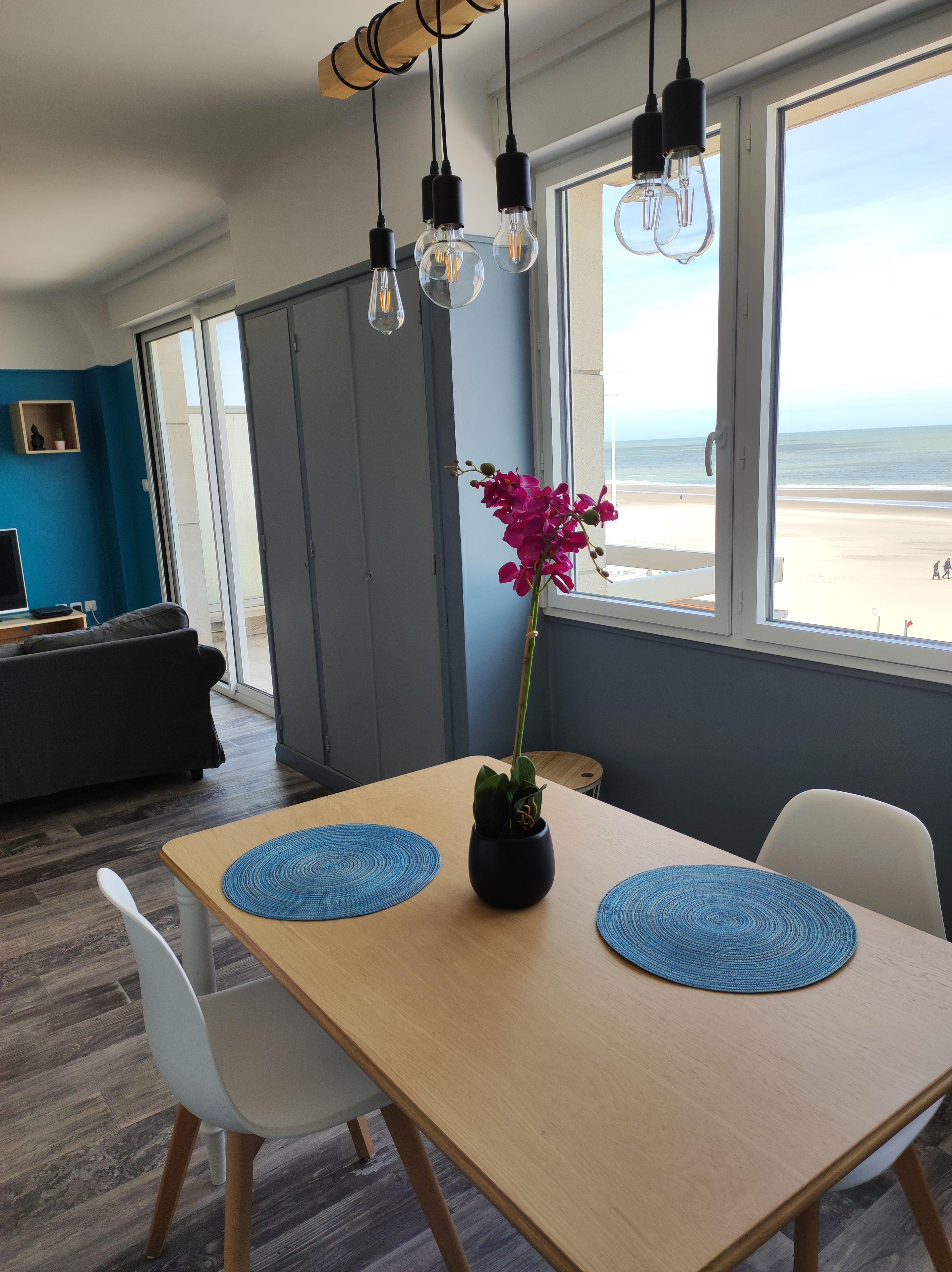 Een moderne eetruimte met een houten tafel, twee witte stoelen, een vaas met een roze bloem en uitzicht op het strand door grote ramen.