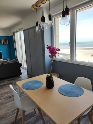 Een moderne eetruimte met een houten tafel, twee witte stoelen, een vaas met een roze bloem en uitzicht op het strand door grote ramen.