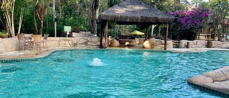 A swimming pool with a thatched gazebo, surrounded by rocks and greenery.