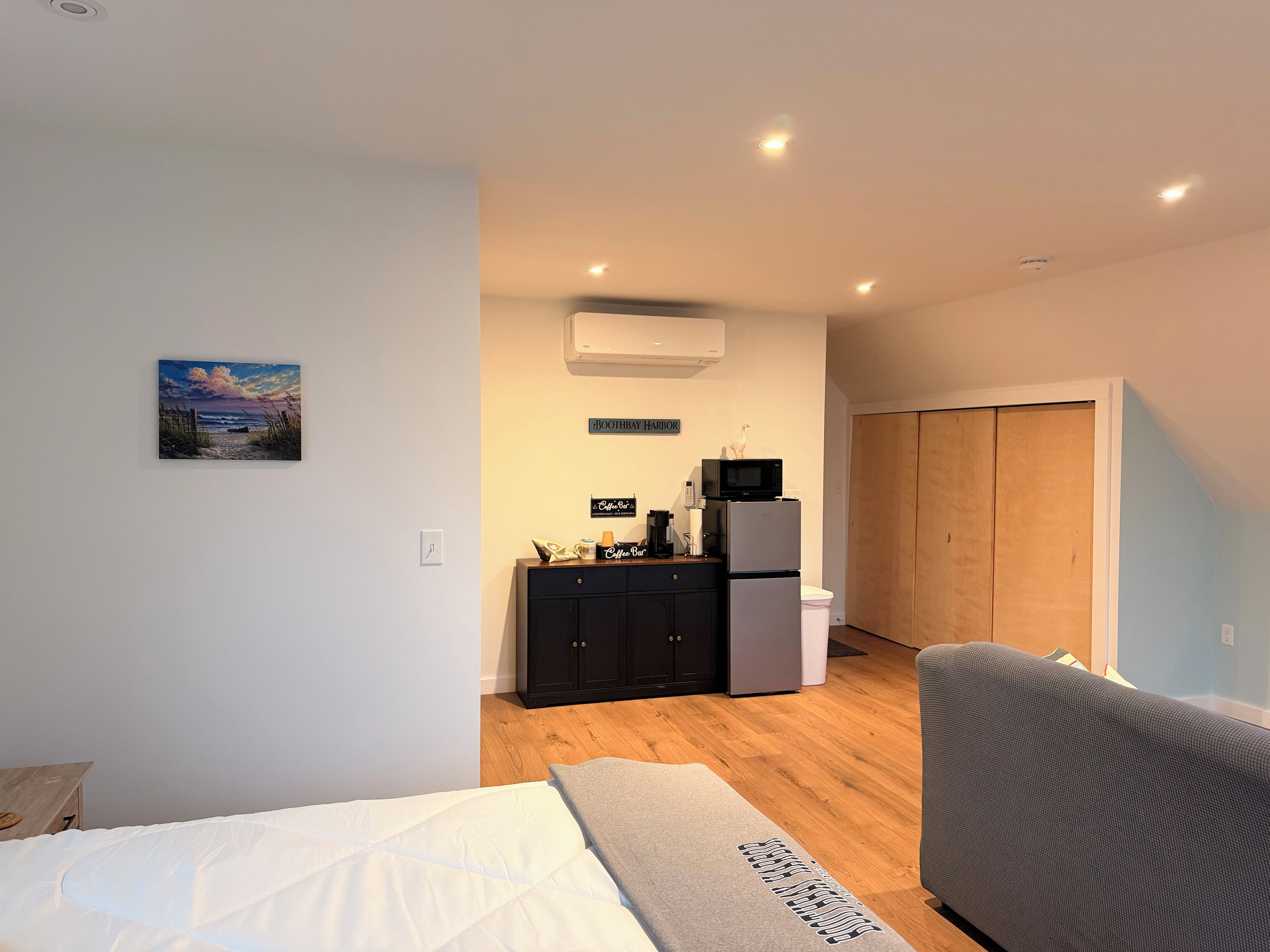 A bedroom with a bed, a grey sofa, a kitchenette with a microwave and refrigerator, and a wooden door.