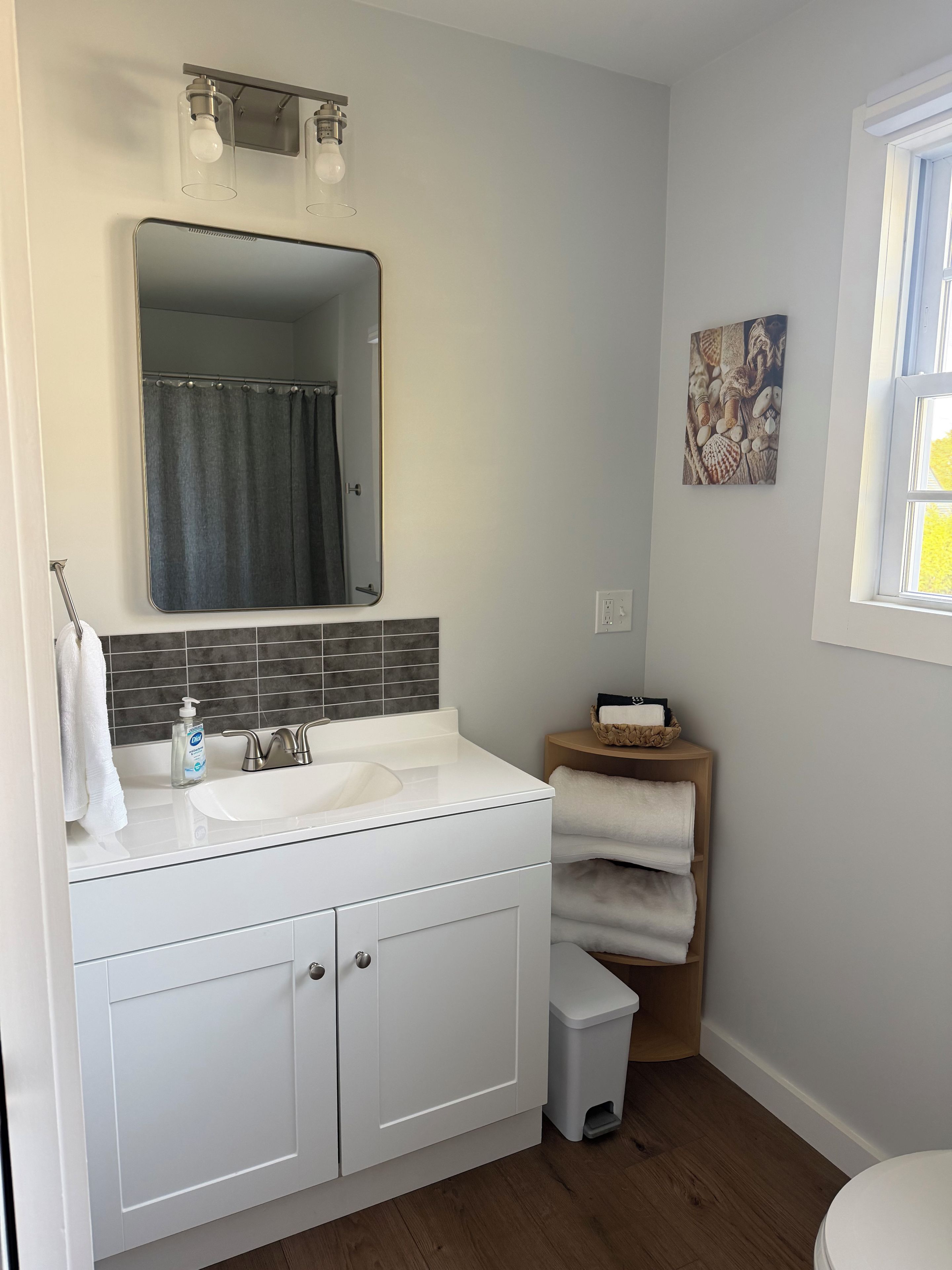A bathroom with a white sink, mirror, and tiled backsplash. There is a small trash bin and a wooden side table with towels.