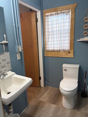 Une salle de bain avec un lavabo blanc, des toilettes, une fenêtre avec un rideau à carreaux et une porte en bois.