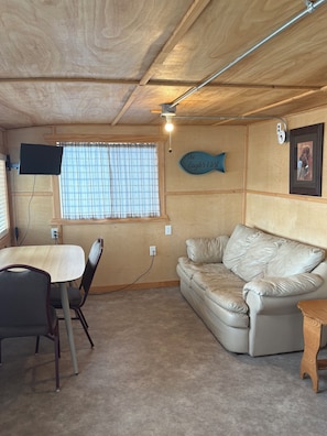 Une pièce avec un plafond en bois, un téléviseur à écran plat fixé au mur, une fenêtre avec des stores, un canapé en cuir beige, une table à manger avec des chaises et une petite table d’appoint en bois.