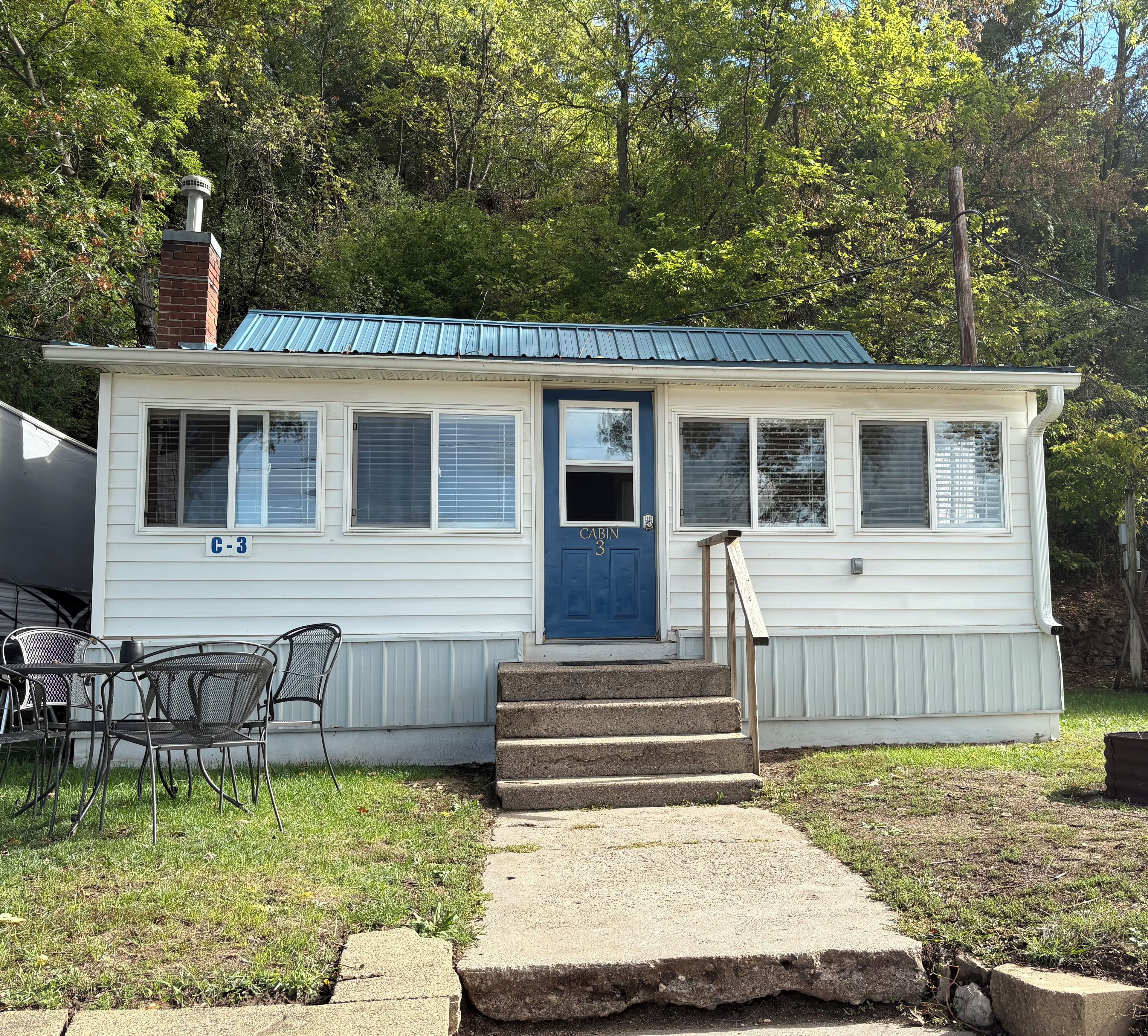 A single-story cabin with a blue door, numbered 'C-3' on the side, a chimney, and a small porch with steps leading to the door.