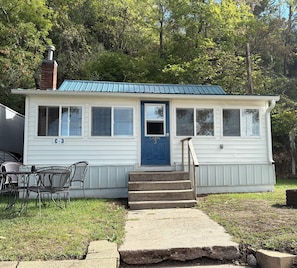 Une cabane de plain-pied avec une porte bleue, portant le numéro « C-3 » sur le côté, une cheminée et un petit porche avec des marches menant à la porte.